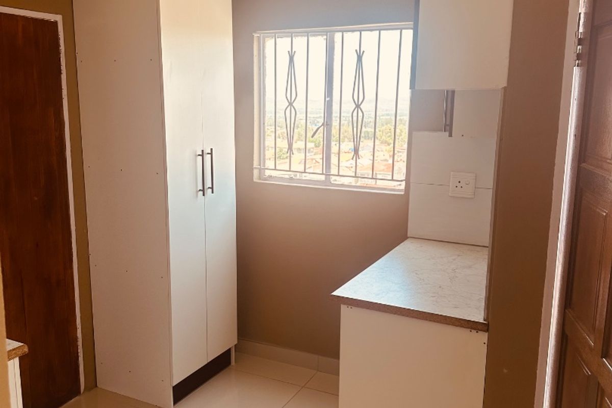 A compact kitchen area featuring white built-in cabinetry, laminate countertops, tiled flooring, and a window with security bars.