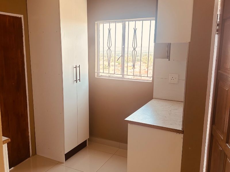 A compact kitchen area featuring white built-in cabinetry, laminate countertops, tiled flooring, and a window with security bars.