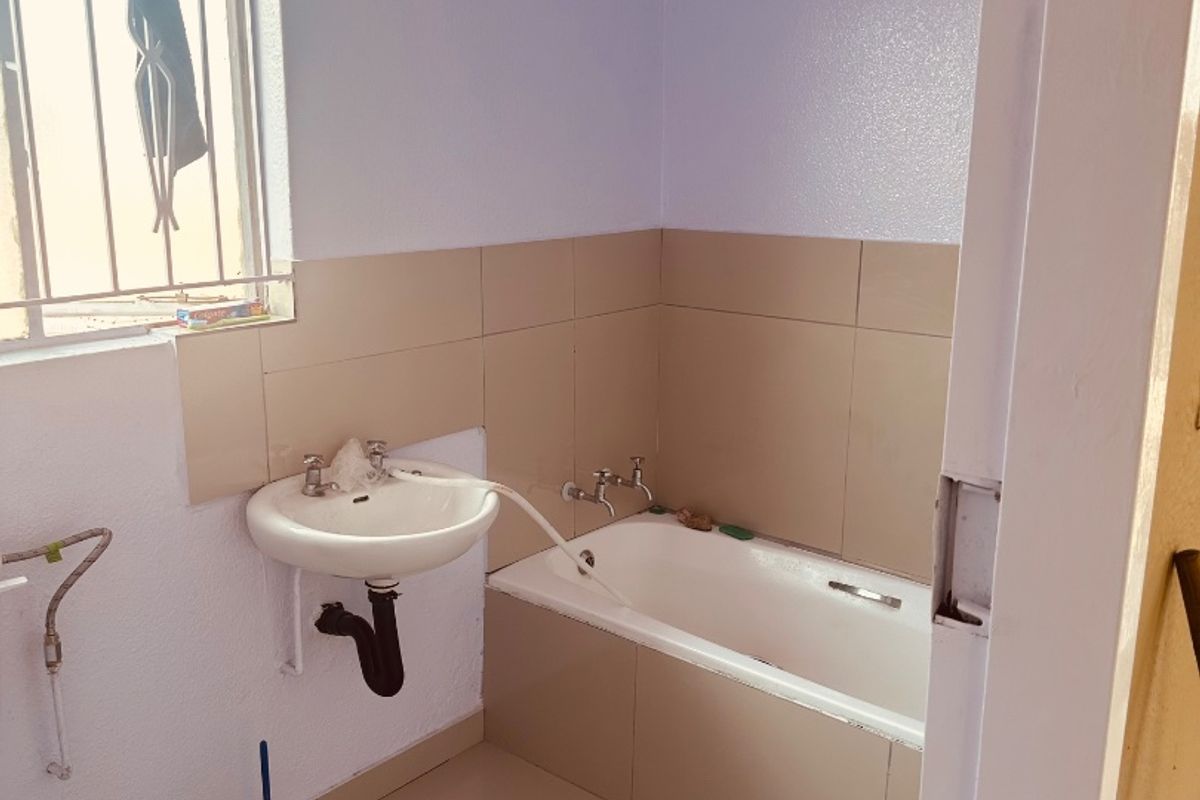 A functional bathroom featuring a bathtub, wall-mounted basin, and neutral wall and floor tiling, with security bars installed on the window.