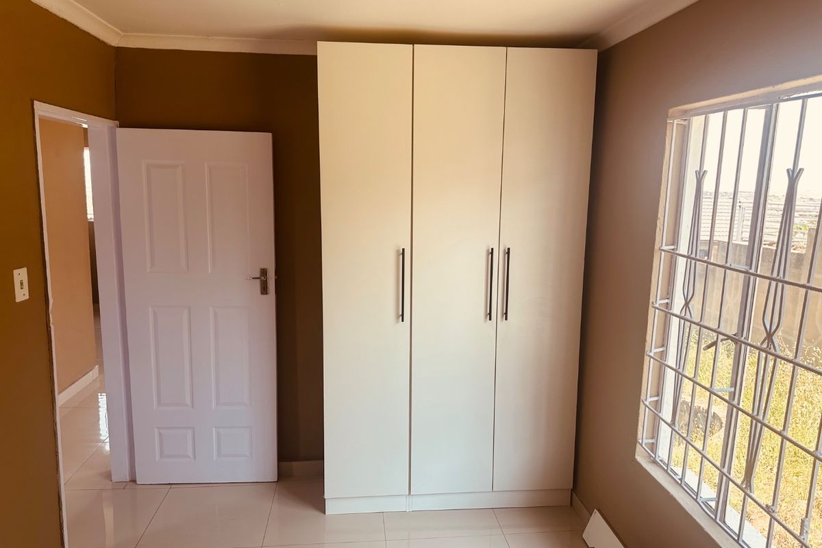 A clean, empty bedroom featuring neutral wall tones, tiled flooring, and a modern built-in wardrobe with security bars on the window.