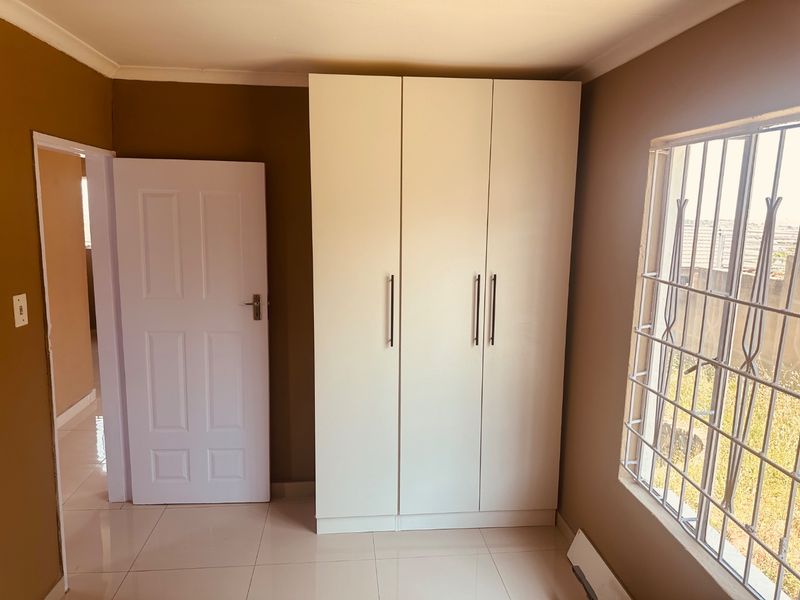 A clean, empty bedroom featuring neutral wall tones, tiled flooring, and a modern built-in wardrobe with security bars on the window.