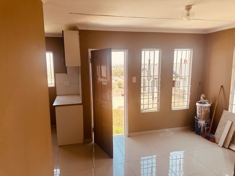 An open-plan living area featuring tiled flooring, security bars on windows, and a small built-in kitchen unit. The space is vacant and requires minor repairs.