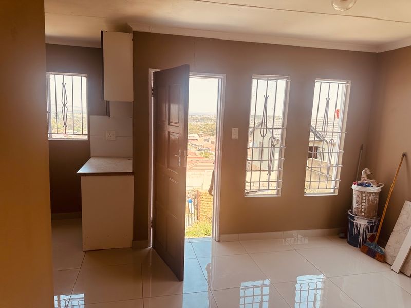 An open-plan living area featuring tiled flooring, security bars on windows, and a compact built-in kitchen unit with direct access to the exterior.