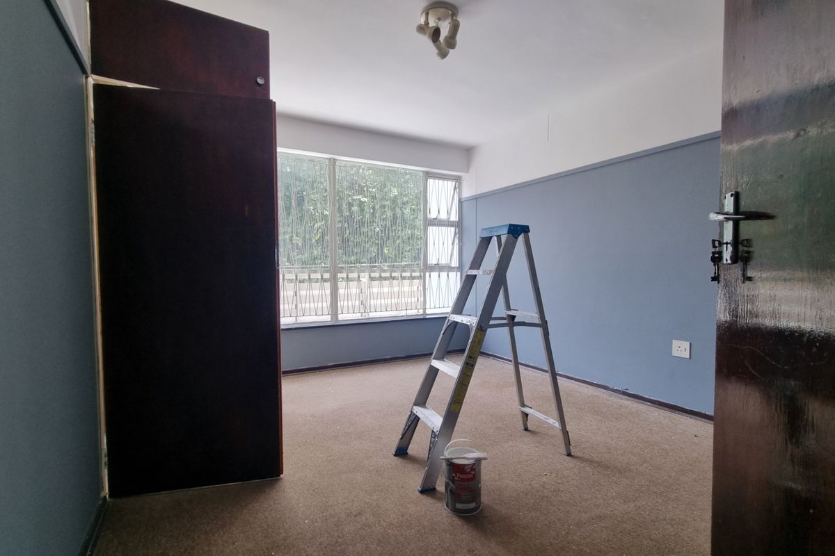 An empty bedroom featuring built-in cupboards, large windows with security bars, and carpeted flooring, currently undergoing minor maintenance.