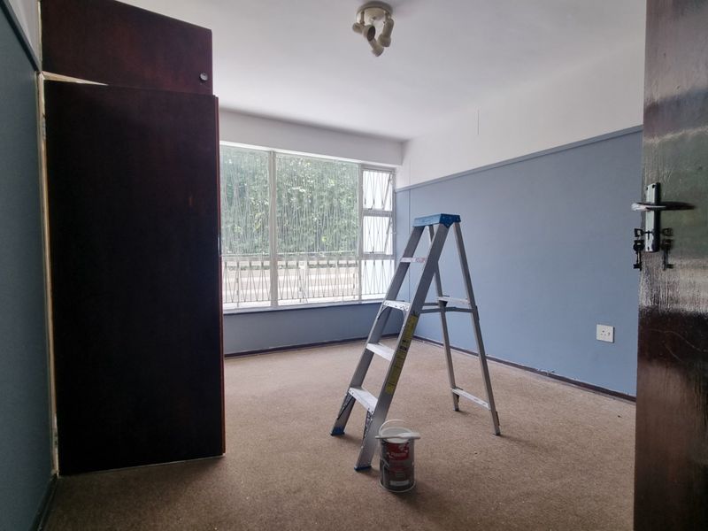 An empty bedroom featuring built-in cupboards, large windows with security bars, and carpeted flooring, currently undergoing minor maintenance.