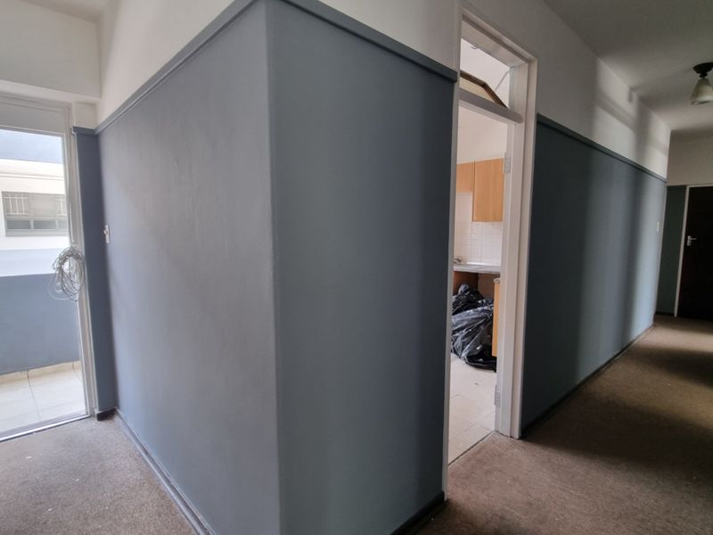 A vacant hallway featuring grey painted walls with dado rails, carpeted flooring, and access to an adjacent balcony and kitchen area.