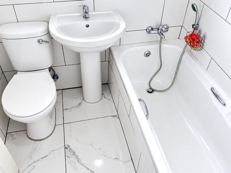 A clean, modern bathroom featuring white wall tiles, marble-effect floor tiles, a pedestal sink, toilet, and a bathtub with a shower mixer.
