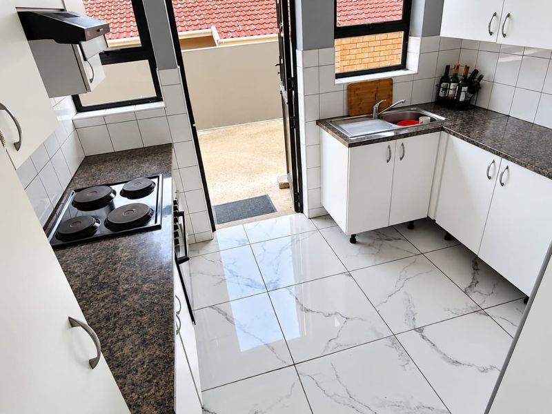 A compact, well-lit kitchen featuring white cabinetry, dark laminate countertops, modern marble-look floor tiles, and direct access to an outdoor area.