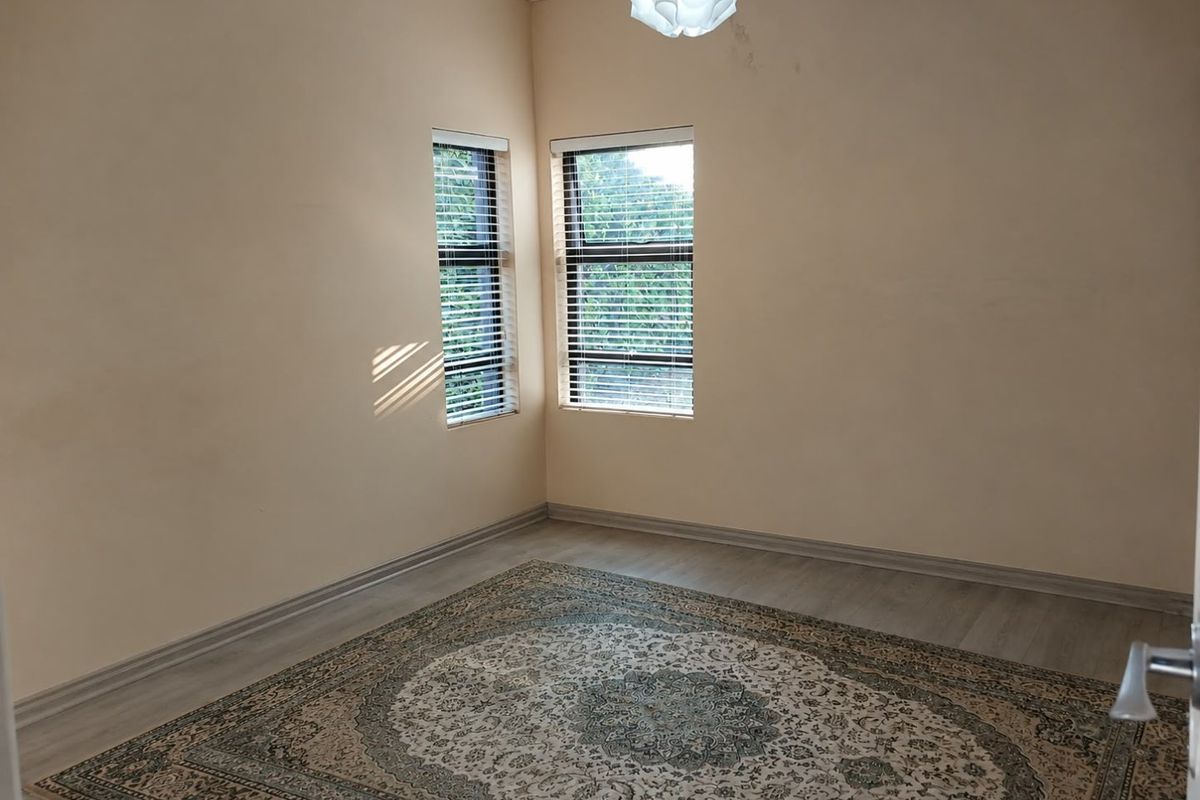 A bright, empty bedroom featuring light-toned laminate flooring, neutral walls, and dual windows fitted with venetian blinds.