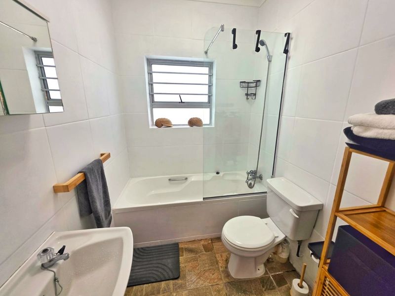 A functional bathroom featuring a bathtub with a glass shower screen, pedestal basin, and toilet, finished with white wall tiles and textured floor tiles.
