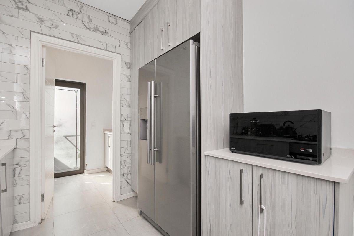 Modern kitchen area featuring light wood-grain cabinetry, stone countertops, and elegant marble-look wall tiling, leading through to a bright utility space.