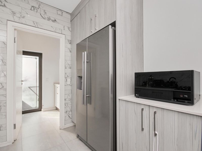 Modern kitchen area featuring light wood-grain cabinetry, stone countertops, and elegant marble-look wall tiling, leading through to a bright utility space.