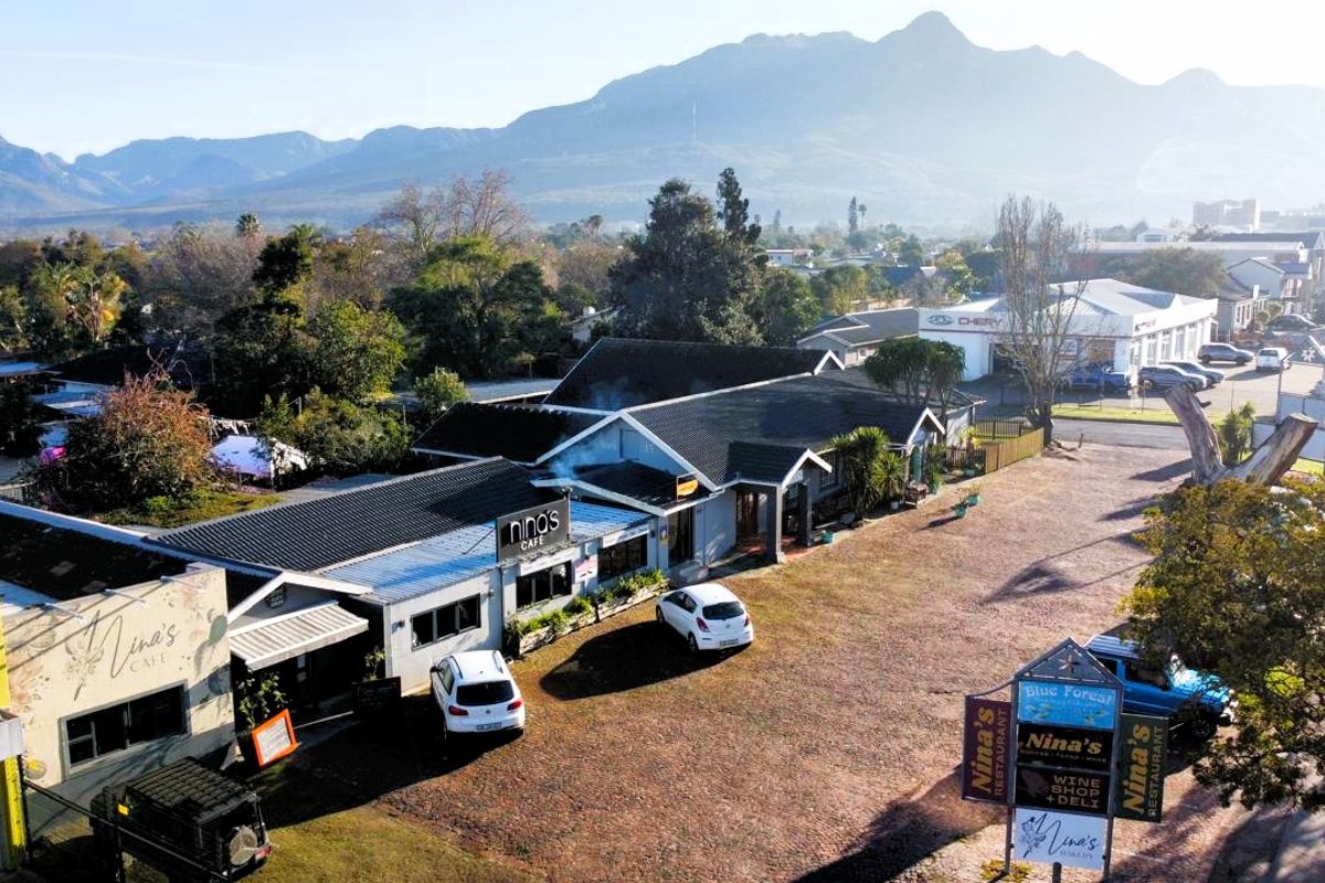 Aerial view of a commercial property featuring a paved parking area, multiple building structures with tiled roofs, and prominent signage against a mountain backdrop.