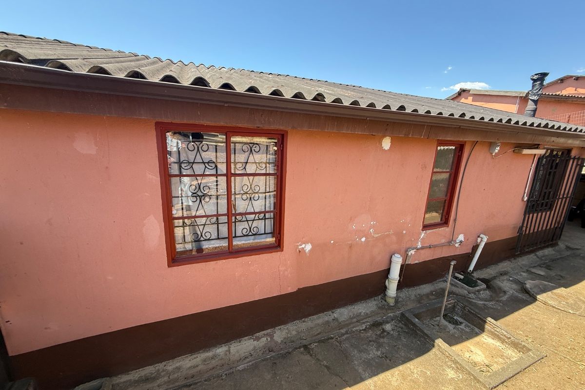 Rear exterior view of the house featuring a corrugated roof, security-barred windows, and exposed external plumbing against a painted masonry wall.