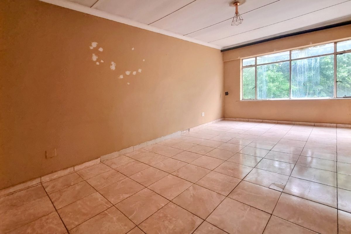 An empty living room featuring tiled flooring, a large window, and a ceiling light fixture, requiring cosmetic updates to the walls and flooring.