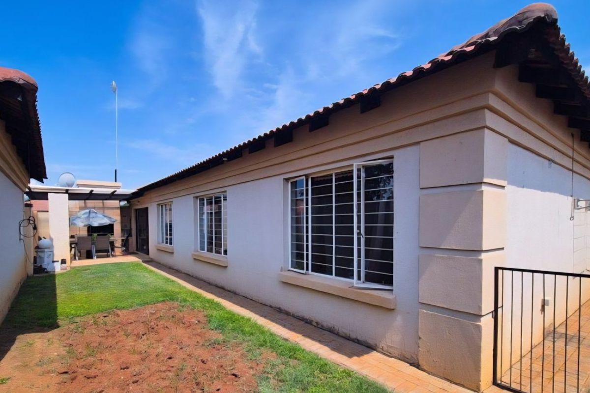 Rear view of the townhouse featuring a paved walkway, security-barred windows, terracotta roof tiles, and a covered outdoor braai area.