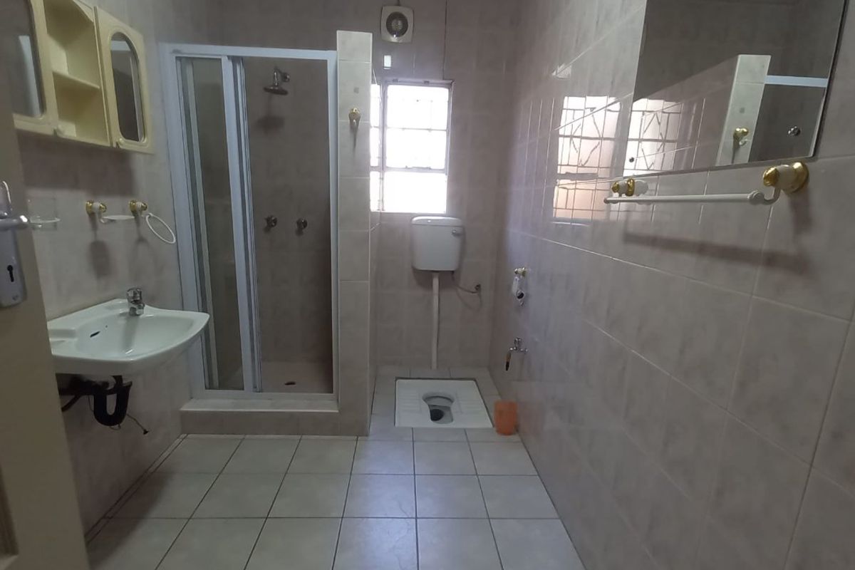 A functional bathroom featuring a shower enclosure, pedestal basin, and a squat toilet with wall-mounted cistern, finished with neutral wall and floor tiles.