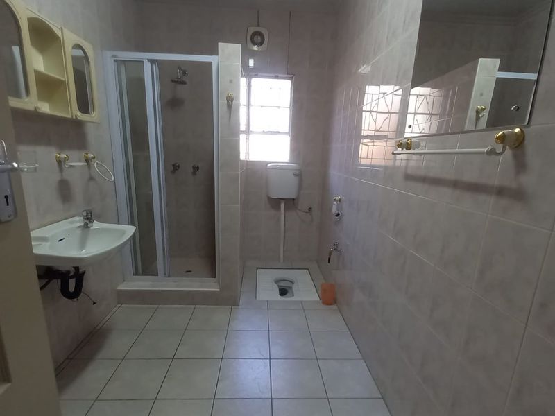 A functional bathroom featuring a shower enclosure, pedestal basin, and a squat toilet with wall-mounted cistern, finished with neutral wall and floor tiles.