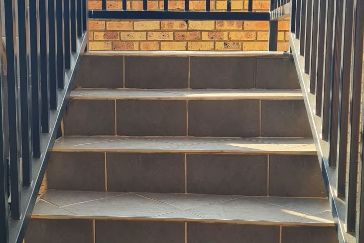 An exterior staircase featuring dark tiled steps and a black metal balustrade, leading up towards a brick boundary wall.