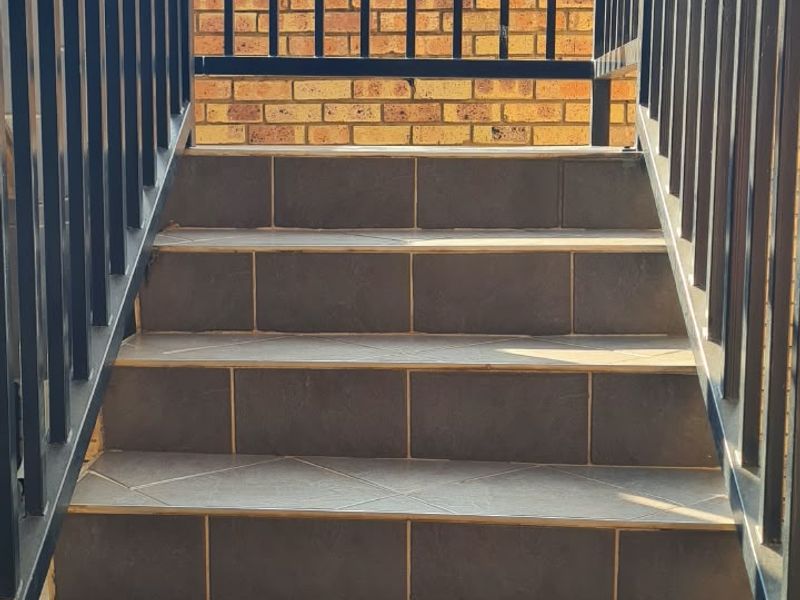 An exterior staircase featuring dark tiled steps and a black metal balustrade, leading up towards a brick boundary wall.