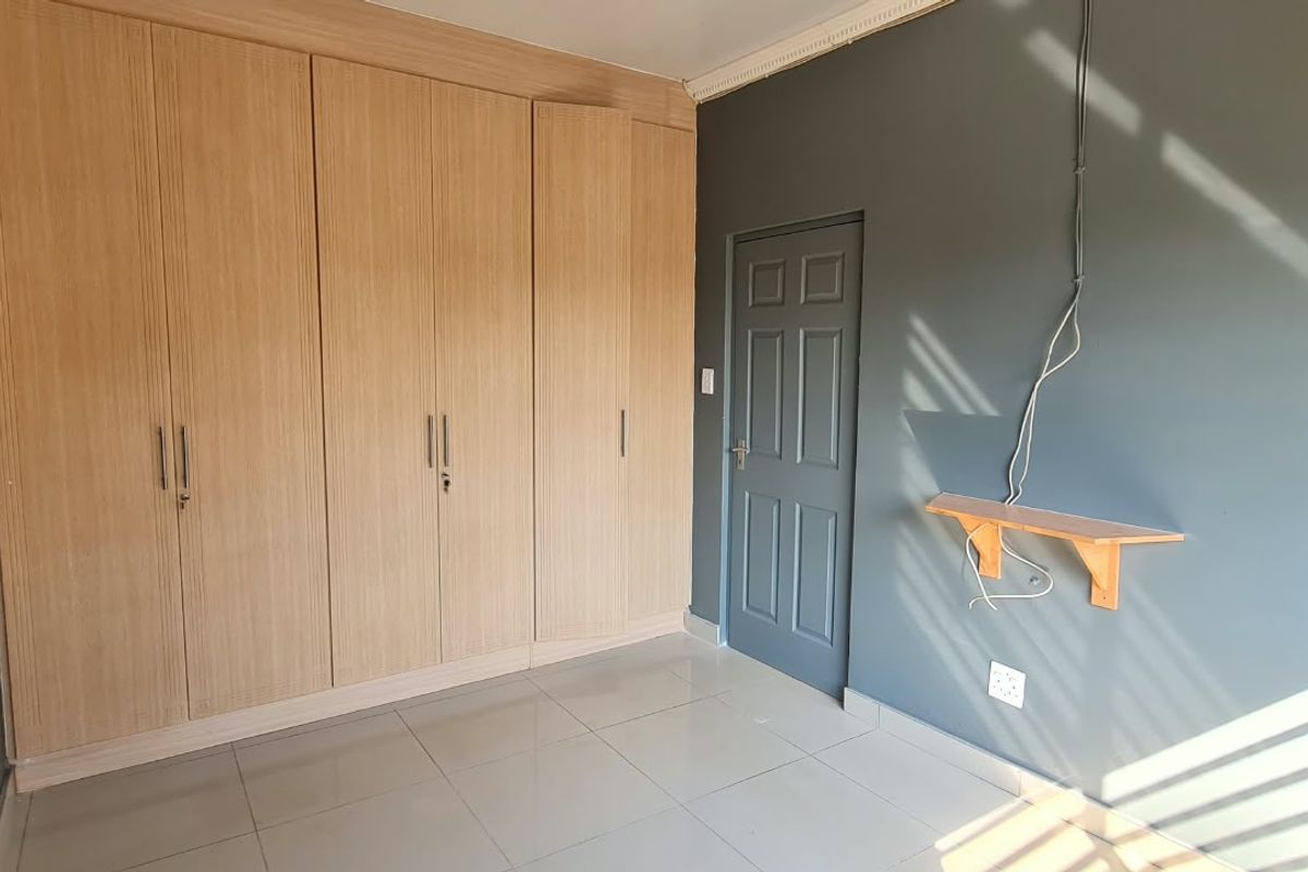 A bright, empty bedroom featuring light wood-finish built-in cupboards, tiled flooring, and dark grey painted walls.