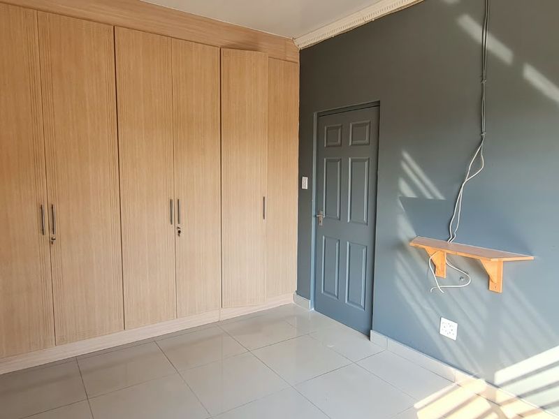 A bright, empty bedroom featuring light wood-finish built-in cupboards, tiled flooring, and dark grey painted walls.