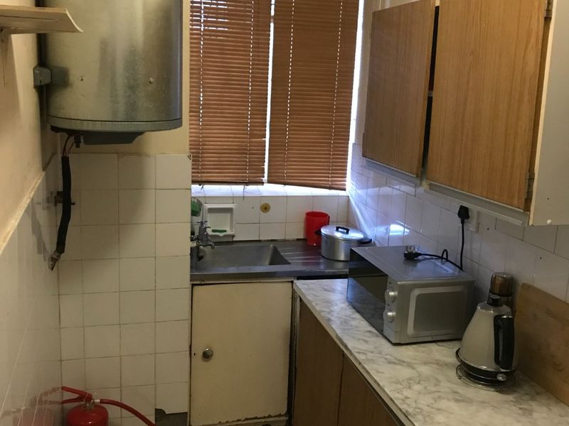 A compact, dated kitchen featuring original cabinetry, a stainless steel sink, tiled splashbacks, and a wall-mounted electric water heater.