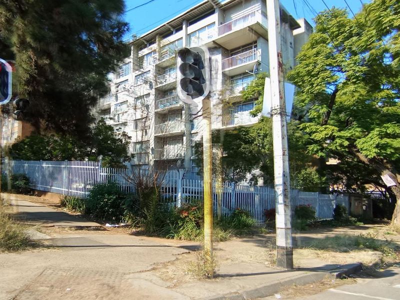 Street view of a multi-story apartment building featuring balconies and perimeter fencing, situated in an established neighborhood with mature trees.