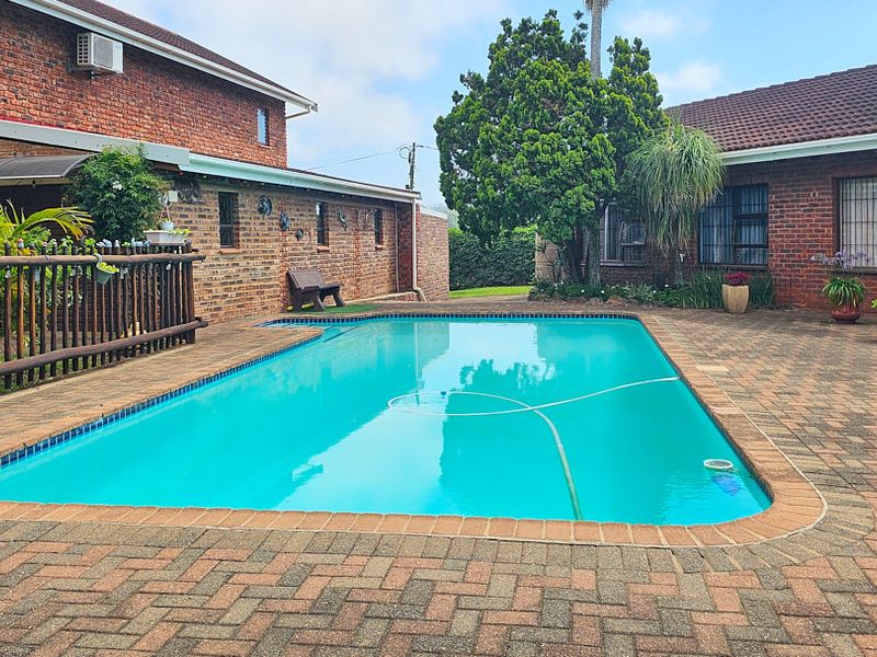A well-maintained outdoor swimming pool area featuring brick paving, surrounded by brick exterior walls and established garden greenery.