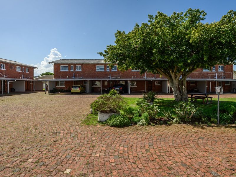 Front view of a brick townhouse complex featuring a paved driveway, covered carports, and established garden area.
