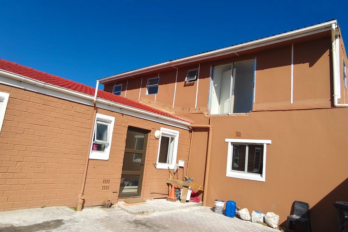 Rear exterior view of a two-story house featuring a paved courtyard, red metal roofing, and multiple windows with white frames.