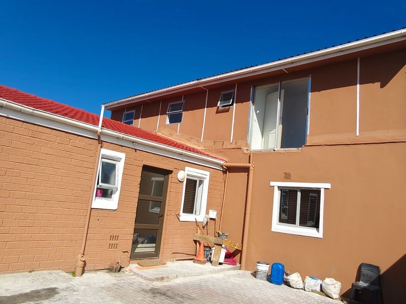 Rear exterior view of a two-story house featuring a paved courtyard, red metal roofing, and multiple windows with white frames.