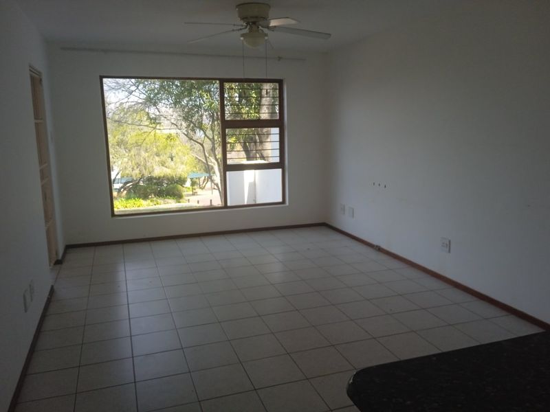An empty living room featuring tiled flooring, a ceiling fan, and a large window providing natural light and garden views.