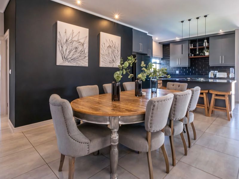 Modern open-plan dining and kitchen area featuring dark cabinetry, a kitchen island with breakfast bar, subway tile backsplash, and contemporary lighting fixtures.