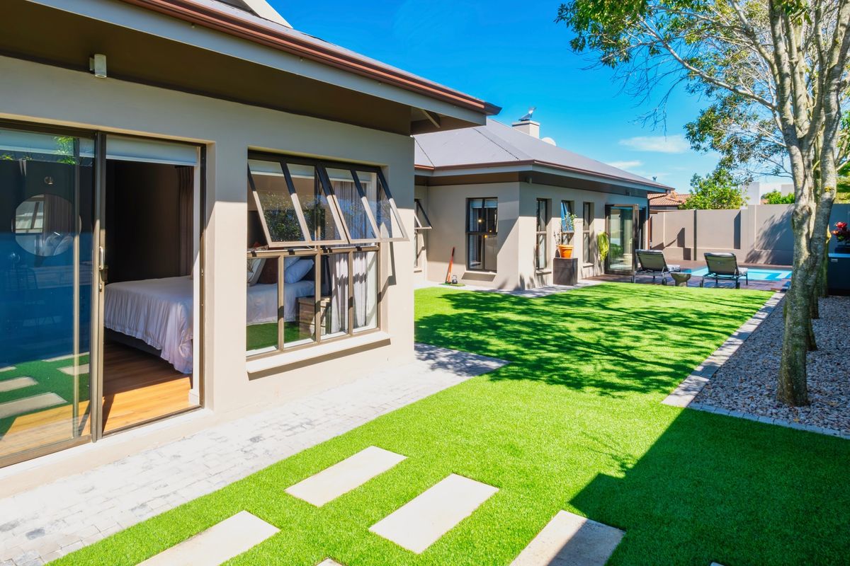 A well-maintained rear exterior view featuring a landscaped artificial lawn, paved pathways, sliding door access to the interior, and a private swimming pool area.