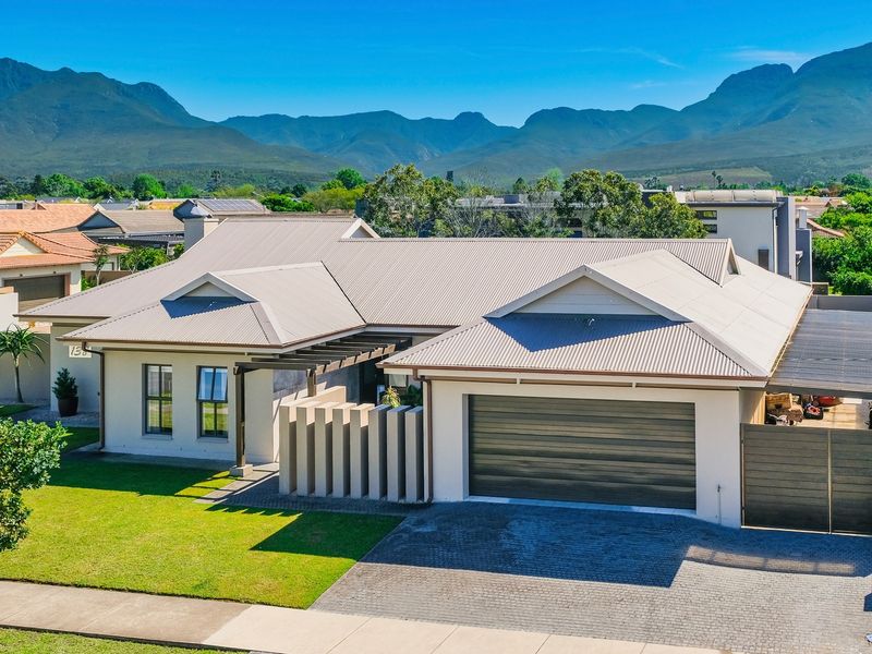 A well-maintained single-story house featuring a modern corrugated metal roof, double garage, paved driveway, and a scenic mountain backdrop.