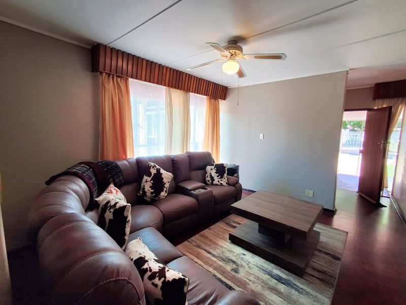 A comfortable living room featuring laminate flooring, a ceiling fan, and a wooden pelmet above the window, leading to an adjacent entryway.