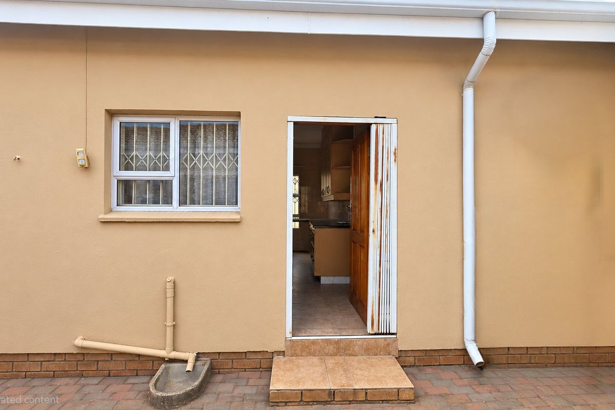 Rear exterior view of the apartment featuring a paved patio area, security gate, and window bars, showing signs of weathering and wear.
