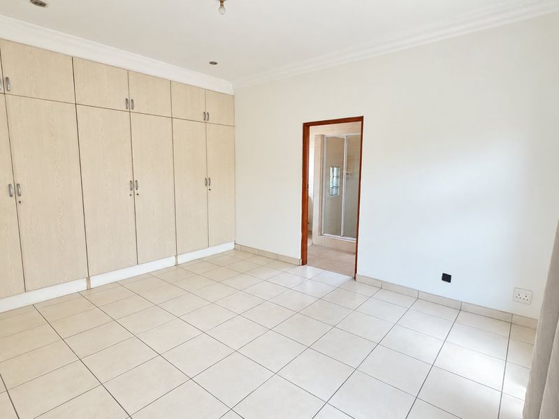 A bright, empty bedroom featuring extensive built-in cupboards, neutral tiled flooring, and direct access to an en-suite bathroom.