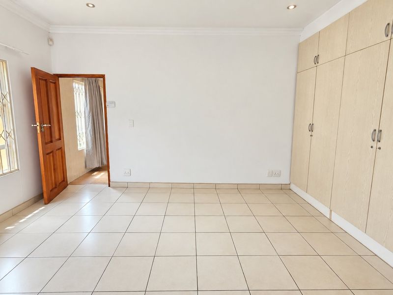 A bright, empty bedroom featuring neutral tiled flooring, ample built-in cupboard storage, and a secure window with a metal security gate.