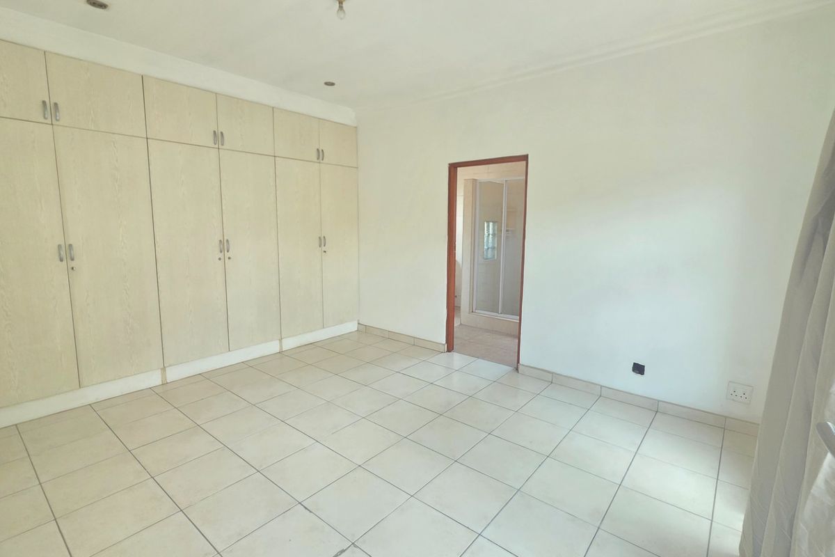A spacious, empty bedroom featuring neutral tiled flooring and extensive built-in cupboard storage, with direct access to an en-suite bathroom.