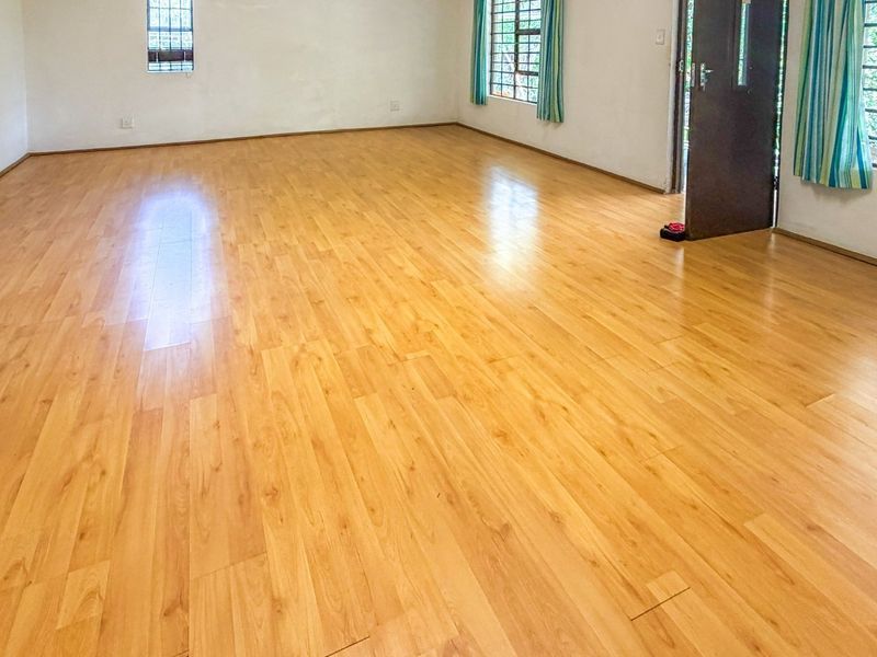 A spacious, empty living area featuring light-toned laminate flooring, large windows with security bars, and neutral wall finishes.