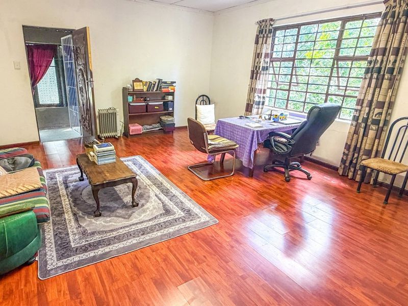 A spacious living area featuring warm-toned laminate flooring, a large window with security bars, and direct access to an adjacent room via a security gate.