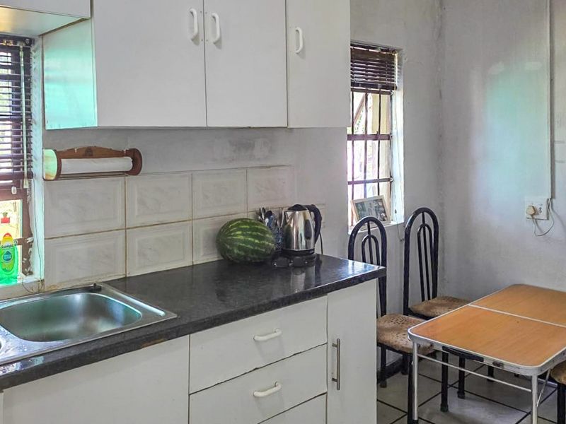 A compact kitchen featuring white built-in cabinetry, a double basin stainless steel sink, tiled backsplash, and security bars on the windows.