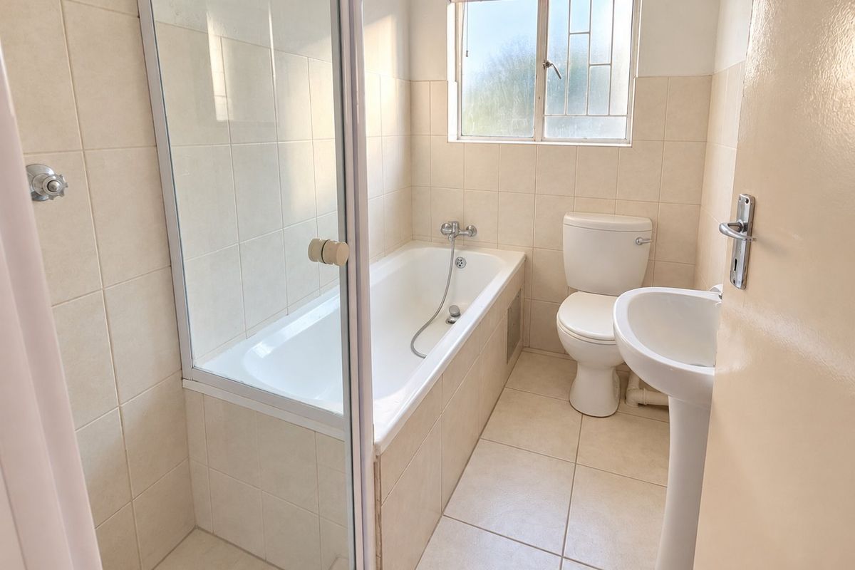 A functional bathroom featuring a built-in bathtub with a glass shower screen, a pedestal washbasin, and a toilet, finished with neutral wall and floor tiles.