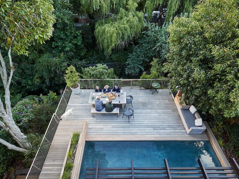 An elevated aerial view of a timber deck featuring a swimming pool, built-in bench seating, and a pergola, surrounded by lush mature trees.
