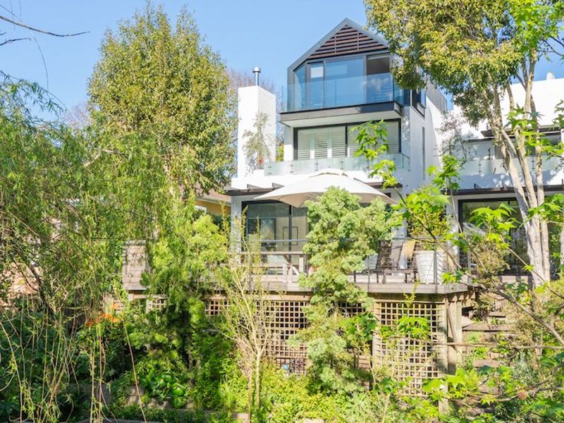 Rear view of a multi-level modern house featuring timber decking, glass balustrades, and tiered garden landscaping with mature trees.