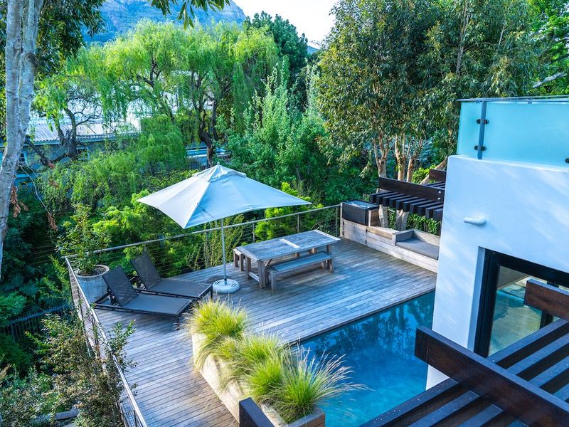 An elevated outdoor deck area featuring a swimming pool, timber decking, glass balustrades, and a pergola, set against a lush, leafy backdrop.