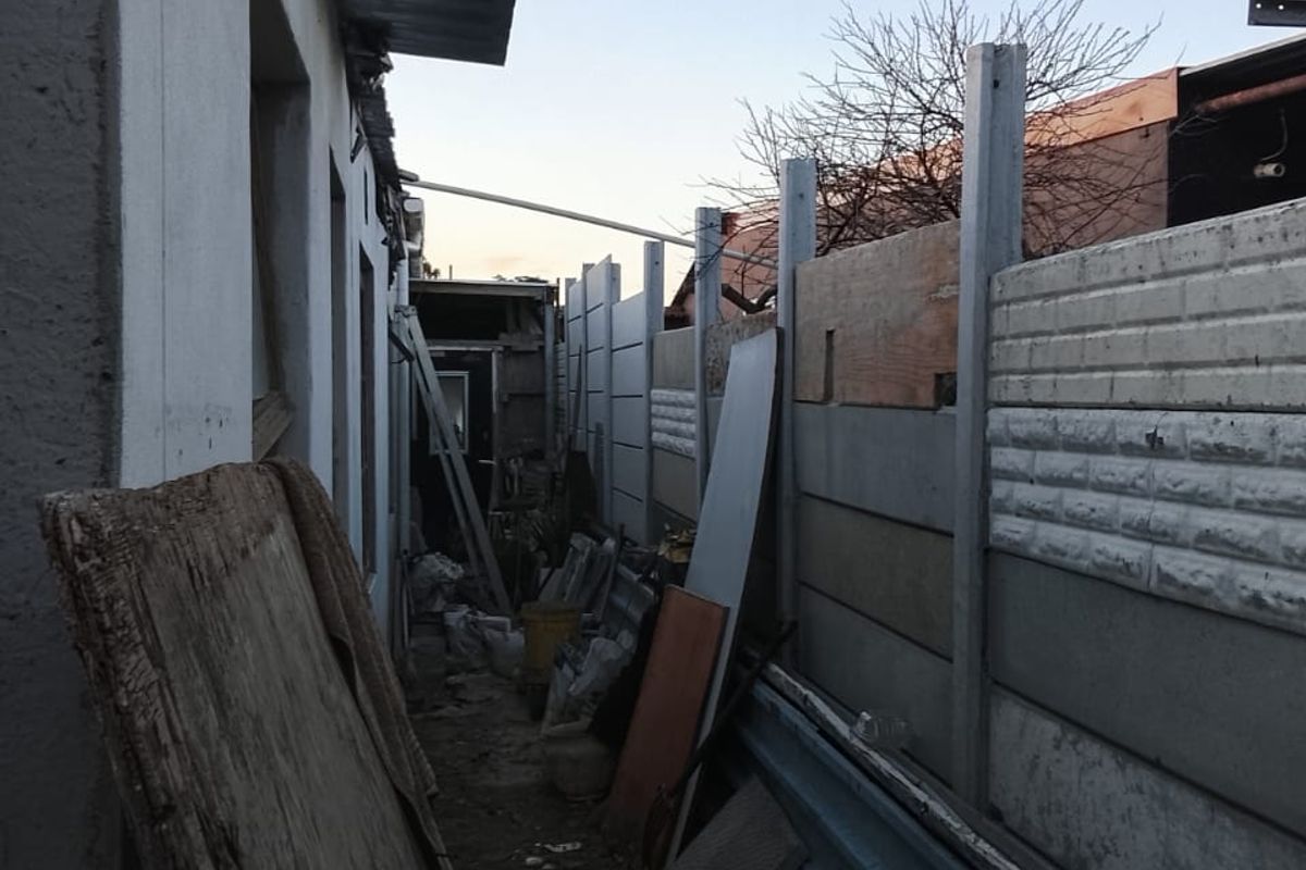A narrow side or rear exterior passage featuring a precast concrete boundary wall and exposed building structure requiring significant maintenance and clearing.