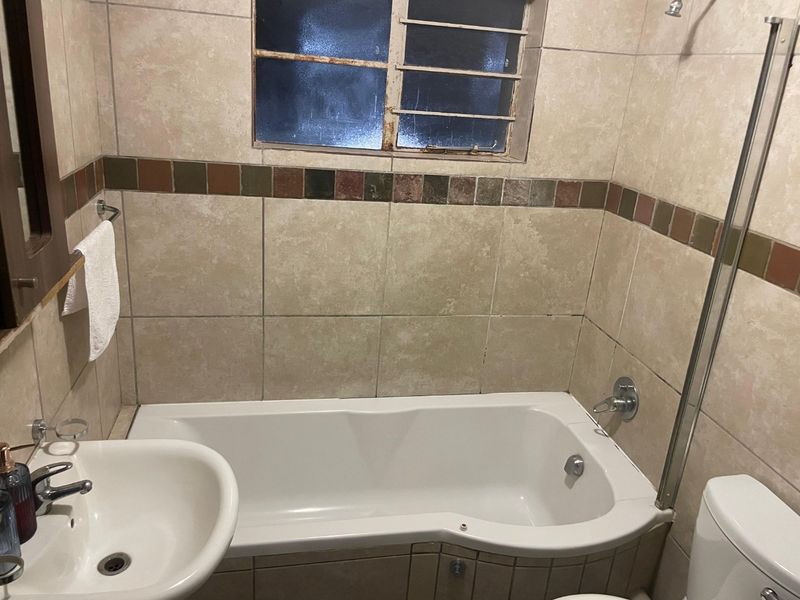 A functional bathroom featuring a bathtub, pedestal basin, and toilet with neutral wall tiling and a window with security bars.
